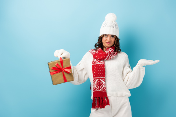 skeptical woman in winter outfit holding wrapped present and gesturing on blue - Foto, Imagem