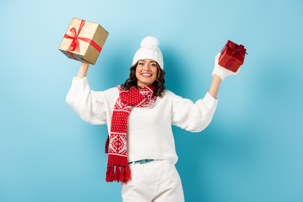 mladá potěšená žena v zimním oblečení držení dárky na modré - Fotografie, Obrázek