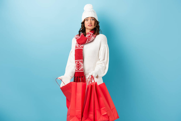 donna sorpresa in abito invernale che tiene borse della spesa rosse su blu - Foto, immagini