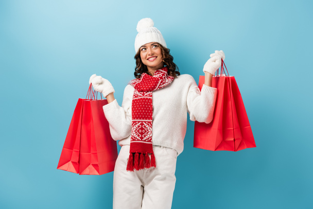 joyful young woman in winter outfit holding red shopping bags and looking up on blue - Photo, Image