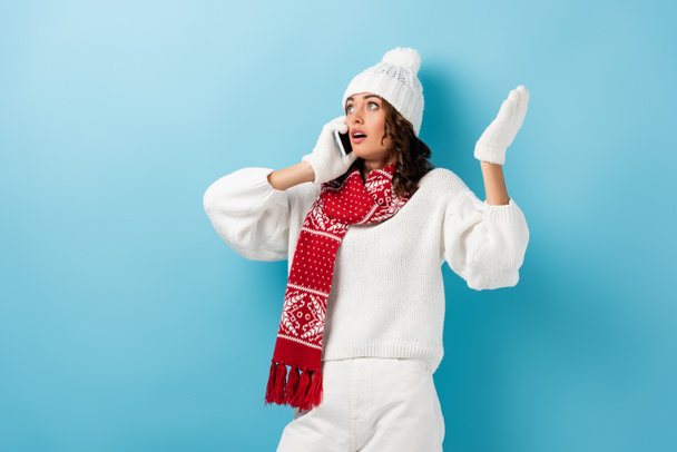 shocked young woman in white winter outfit talking on smartphone and gesturing on blue - Photo, Image