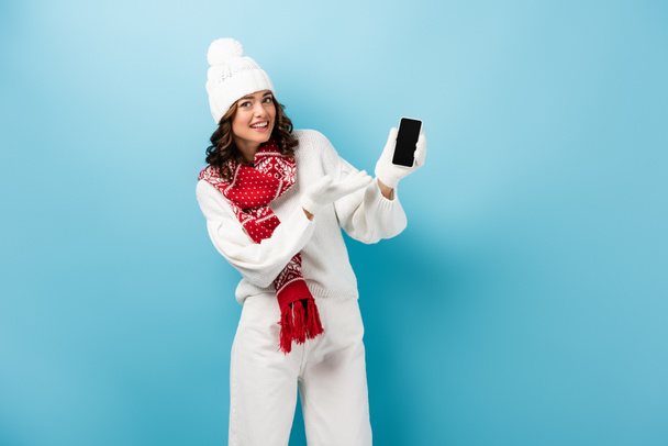 young woman in winter outfit pointing with hand at mobile phone with blank screen on blue  - Photo, Image
