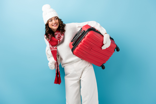 joyful young woman in winter outfit carrying red suitcase on blue - Фото, зображення