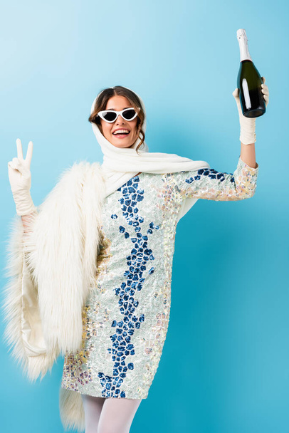 mulher elegante em óculos de sol sorrindo enquanto segurando garrafa de champanhe e mostrando sinal de paz no azul - Foto, Imagem