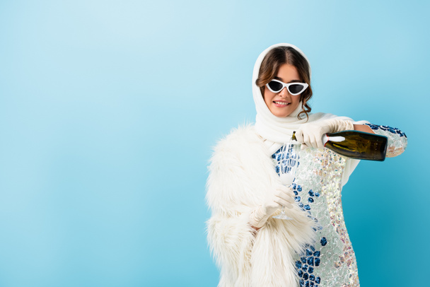 mujer complacida en gafas de sol sosteniendo botella y vertiendo champán en vidrio sobre azul - Foto, imagen