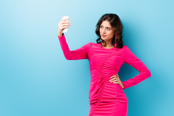brunette woman taking selfie while standing with hand on hip on blue  - Фото, зображення