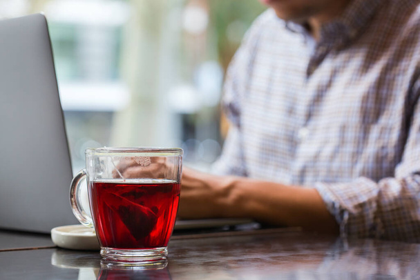 Joven empresario exitoso sentado en un café, bebiendo té rojo saludable y trabajando en un ordenador portátil. Composición de estilo de vida con luz natural  - Foto, imagen