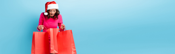 horizontal concept of young excited woman in santa hat looking at shopping bags on blue - Photo, Image