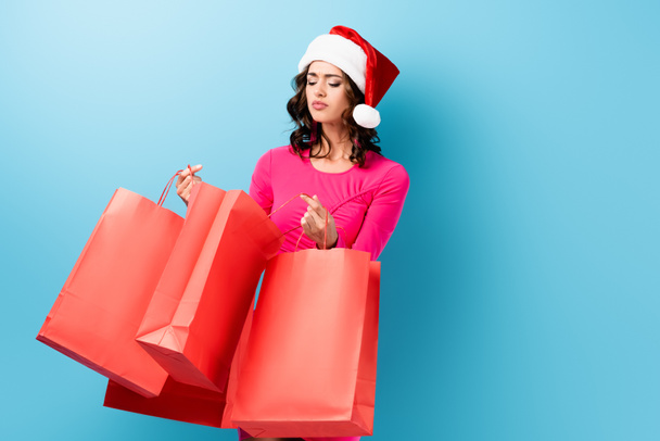 young brunette woman in santa hat looking at shopping bags on blue - Fotografie, Obrázek