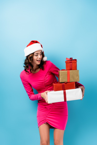 young excited woman in santa hat and dress looking at wrapped presents on blue - Photo, Image