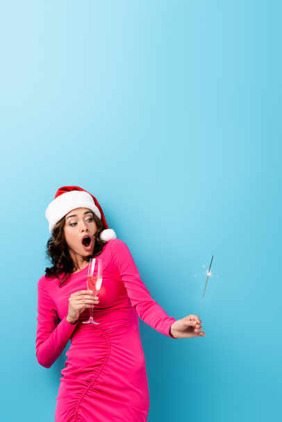 Schockierte Frau mit Weihnachtsmütze mit Wunderkerze und Glas Champagner auf blauem Grund - Foto, Bild