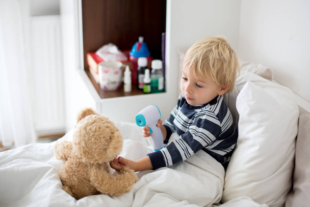 Little toddler blond child, playing with teddy bear in bed, while being sick, checking his temperature with digital thermometer - 写真・画像