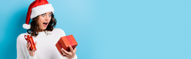 panoramic crop of young shocked woman looking at gift box on blue - Photo, Image