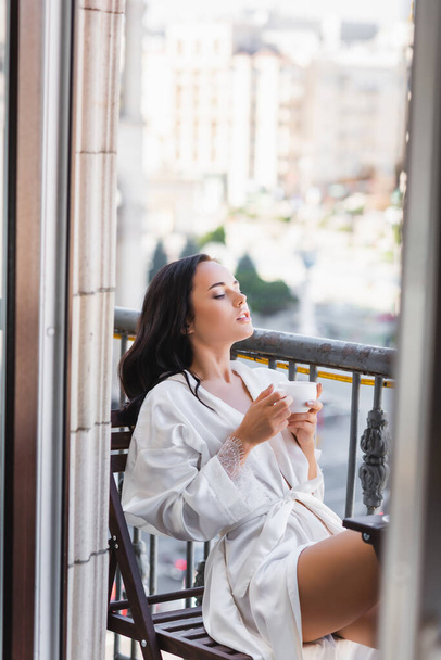 beautiful brunette woman in white robe drinking coffee on balcony - Фото, изображение