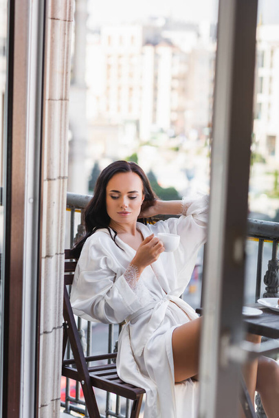 beautiful brunette woman in white robe drinking coffee on balcony - 写真・画像
