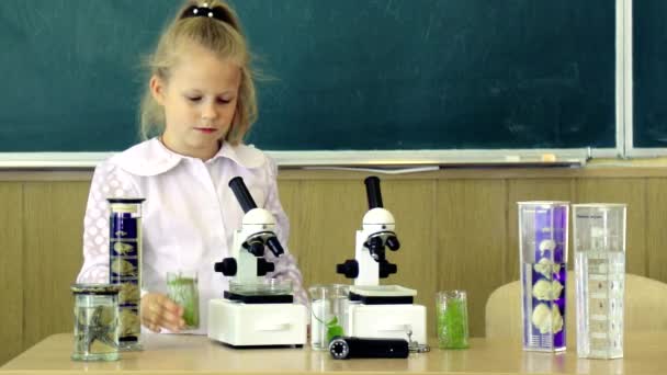 Маленькая девочка возле микроскопа в классе, доска на заднем плане. Сначала интересовался учебой, обучением, образованием. Энтузиастичная концепция ученика. Ребенок, зрачок на спокойном лице возле микроскопа. - Кадры, видео