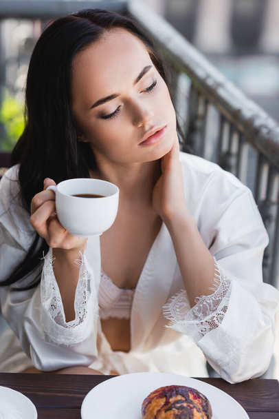brunette woman in white robe drinking coffee and touching neck on balcony - Fotoğraf, Görsel