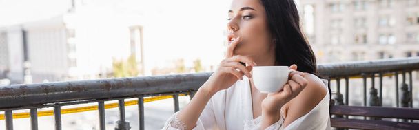 Beyaz bornozlu esmer kadın kahve içiyor ve parmaklarını ağzının yanında tutuyor. - Fotoğraf, Görsel