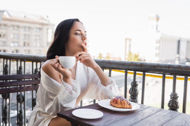 brunetta donna in vestaglia bianca bere caffè e tenendo le dita vicino alla bocca - Foto, immagini