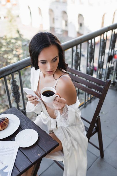 όμορφη μελαχρινή γυναίκα σε λευκό ρόμπα κρατώντας smartphone και πίνοντας καφέ στο μπαλκόνι - Φωτογραφία, εικόνα