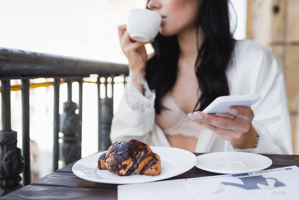 Beyaz bornozlu esmer kadının balkonda kahve içip elinde akıllı telefon tutarken kısmi görüntüsü. - Fotoğraf, Görsel