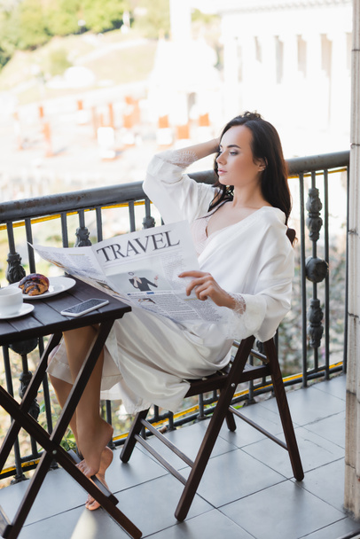 Beyaz cüppeli esmer kadın balkonda oturuyor ve gazete okuyor. - Fotoğraf, Görsel