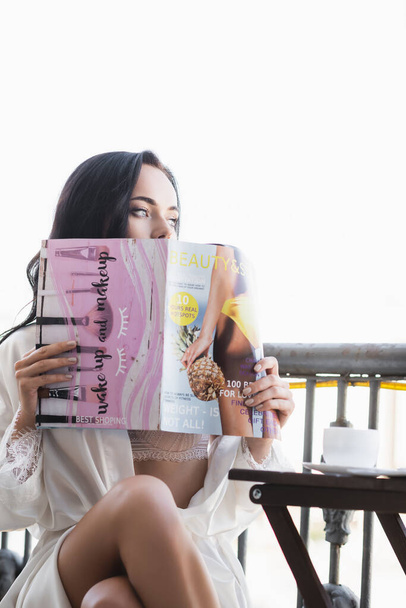 brunette woman in white robe sitting on balcony and reading magazine - Zdjęcie, obraz