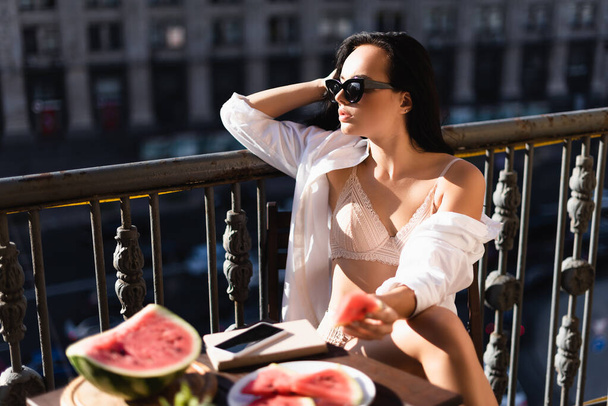 brünette Frau in beiger Unterwäsche und weißem Hemd isst Wassermelone auf Balkon - Foto, Bild