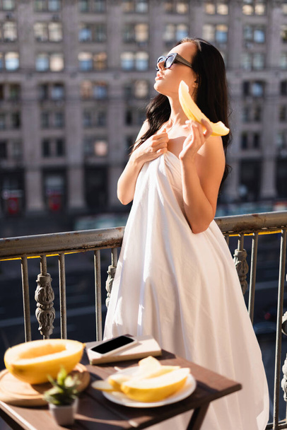 sexy morena mujer en gafas de sol y cubierto de sábana blanca comiendo melón en balcón - Foto, imagen