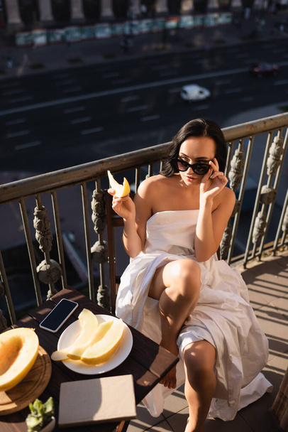 sexy brunette vrouw verhogen zonnebril en eten meloen op balkon - Foto, afbeelding