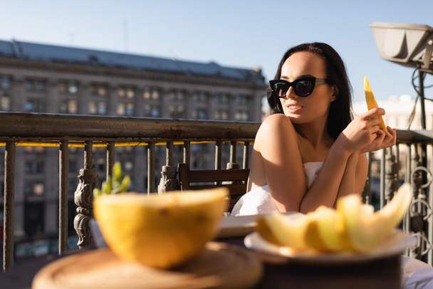 sexy morena mulher em óculos de sol e coberto de branco folha comer melão na varanda - Foto, Imagem