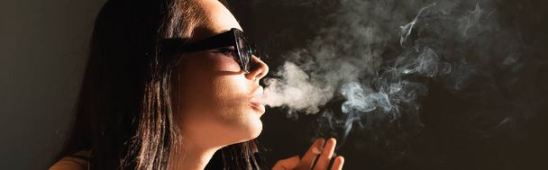 Güneş gözlüklü esmer kadın sigara içiyor, yatay afiş. - Fotoğraf, Görsel