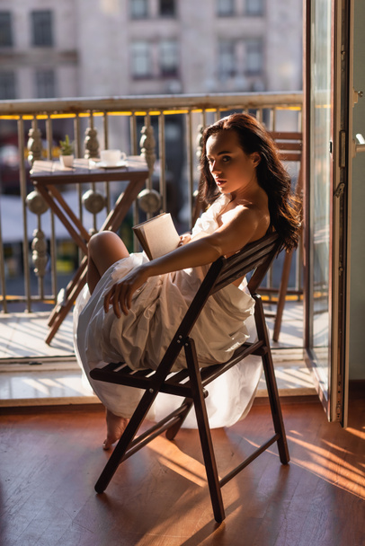 aantrekkelijke vrouw bedekt met wit laken holding boek en terugkijkend - Foto, afbeelding