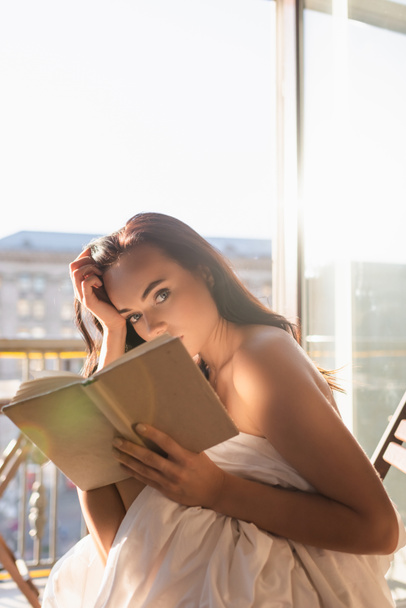 atraktivní žena pokrytá bílým listem čtení knihy a sedí na balkóně - Fotografie, Obrázek