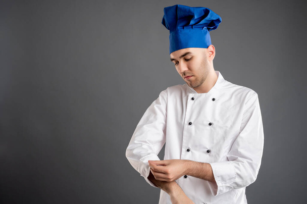 Retrato de hombre joven vestido con un traje de chef blanco organiza su uniforme posando sobre un fondo gris con área de publicidad de espacio de copia - Foto, Imagen