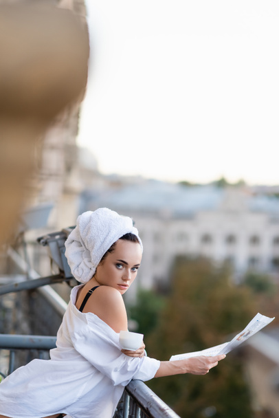 Kafasında havluyla beyaz gömlekli seksi genç kadın elinde gazete tutarak balkonda çay içiyor. - Fotoğraf, Görsel