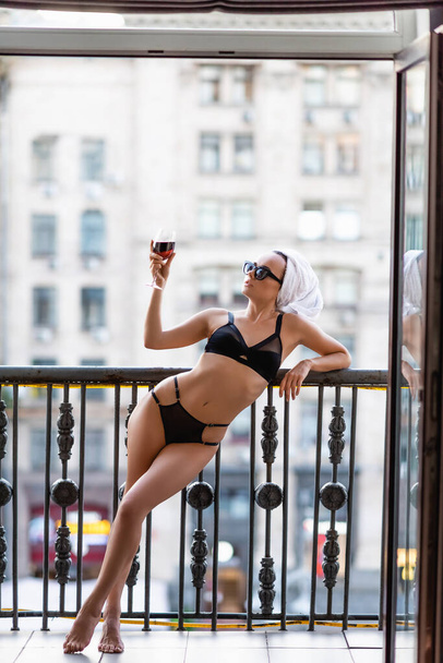 sexy jonge vrouw in zwart ondergoed met handdoek op hoofd drinken rode wijn op balkon - Foto, afbeelding