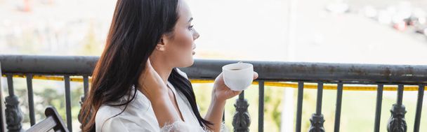 brunette woman in white robe drinking tea and sitting on balcony, horizontal banner - Foto, afbeelding