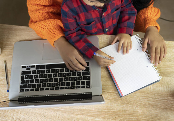 manos de un niño y su madre escribiendo en un cuaderno en un escritorio en casa, con computadora portátil - Foto, Imagen
