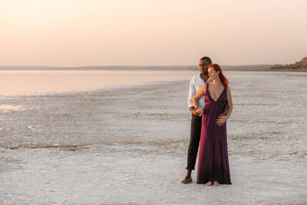 Portret uroczego młodzieńca przytulającego swoją dziewczynę od tyłu stojąc razem. Szczęśliwa mieszana rasa tysiącletnia para ciesząca się dniem na plaży. - Zdjęcie, obraz