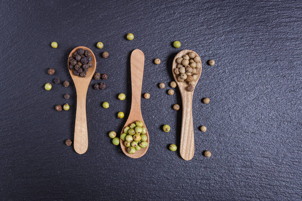 Green pepper, black and whole white peppercorns, pepper mix in wooden spoons on dark background, top view - Fotó, kép