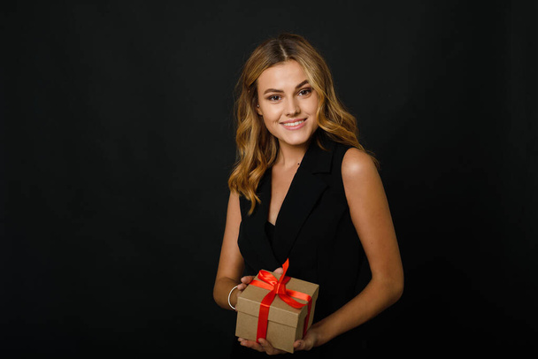 A lovely blonde on a black background is holding a craft gift with a red ribbon. The concept of buying gifts on black friday for christmas and new year - Photo, Image