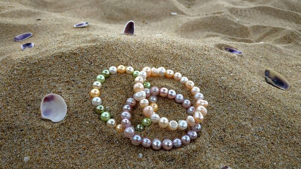 Pulseras iridiscentes multicolores perla se encuentran en la duna de arena amarilla con conchas púrpuras junto al mar. Joyería de perlas naturales vibrantes dan la sensación de lujo rico y armonía.  - Foto, imagen
