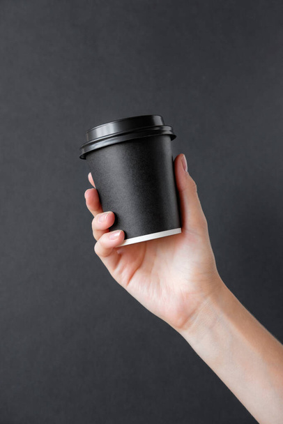 A young girl holds in her hand a black cup of tea or coffee. Mock up coffee or tea paper cup with cap. - Photo, Image