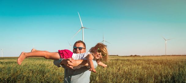 Holčička v otcových rukách před větrnými mlýny. Vtipný kluk lítá. Obnovitelné zdroje energie a udržitelné zdroje. dítě hrající si s tátou u větrné turbíny - Fotografie, Obrázek