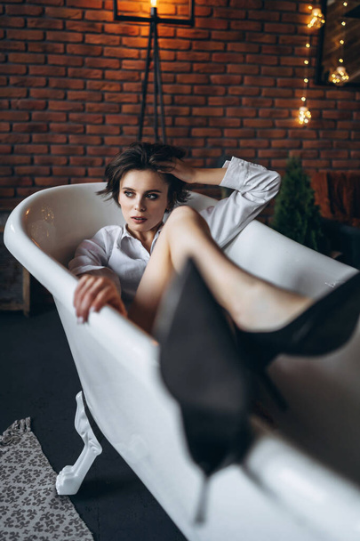 Portrait of a beautiful brunette lying in an empty bathroom holding legs up, wearing a white shirt and black shoes on red brick wall background. - Foto, Bild