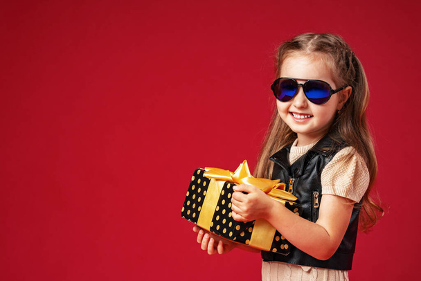 Stylish little girl in sunglasses and a leather vest with a gift box in her hands on a red background. The child is happy and enjoying holiday. Christmas gift. Advertizing. Christmas eve and spirit - Photo, Image