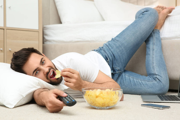 Lazy young man with bowl of chips watching TV while lying on floor at home - Фото, зображення