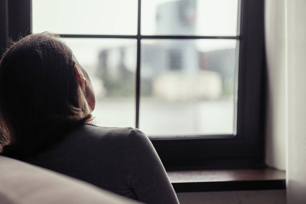 Rückansicht einer einsamen, traurigen, brünetten Frau, die am Fenster sitzt. Konzept - Foto, Bild