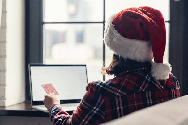 Woman in a red santa claus hat holding credit card using laptop for making order sitting near window. Christmas online shopping. Concept - Valokuva, kuva